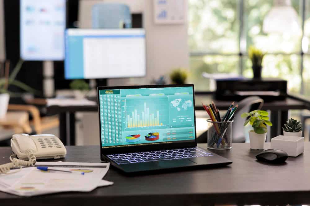 Ein Laptop steht auf einem Schreibtisch. Es werden auf dem Laptop Statistiken angezeigt. Im Hintergrund befindet sich ein Monitor mit weiteren Statistiken.